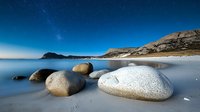 Serene coastal landscape with smooth boulders on sandy beach against mountainous backdrop and deep blue sky, long exposure creates silky water effect.
