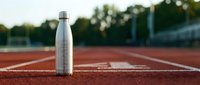 Stainless steel water bottle on running track surface with blurred athletic field background, sports hydration and fitness concept.