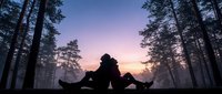 Young adult male in silhouette sitting in wide-legged position on forest path at sunset, surrounded by tall pine trees, purple and orange sky, contemplative mood, suitable for wellness and mindfulness content.