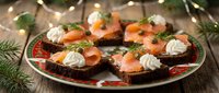 Festive smoked salmon canapes with cream cheese and dill on dark bread, arranged on decorative plate with Christmas lights and pine branches for holiday entertaining.