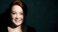 Young Caucasian woman with red hair smiling warmly against dark teal background. Professional headshot portrait with natural lighting and copy space.