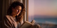Young woman with curly hair reading book by window during sunset, creating peaceful atmosphere for relaxation and mindfulness.