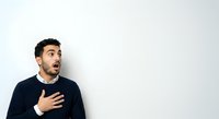 Man with dark hair and beard, appearing surprised or shocked, hand on chest, wearing dark navy sweater against white background. Suitable for expressions, emotions, reactions, advertising, web design.