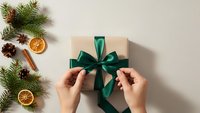 Female hands tying green satin ribbon bow on kraft paper gift box with festive Christmas decorations including pine branches, cones and dried oranges.