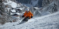 Skier in bright orange jacket carving through fresh powder on mountain slope with alpine village nestled among snow-covered trees in background.