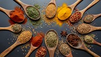 Colorful spices and herbs arranged in wooden spoons on dark background. Cooking ingredients including turmeric, paprika, coriander seeds for culinary recipes.