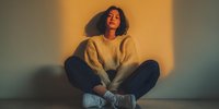 Young Hispanic woman in cozy beige sweater sitting cross-legged on floor with warm sunset lighting creating dramatic shadows on wall behind her.