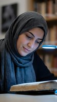 Woman wearing gray hijab reading open book at table with blue lamp in library setting, focused on education and learning.