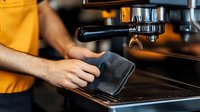 Professional barista in yellow shirt operating commercial espresso machine, cleaning equipment with cloth for coffee shop maintenance and hygiene standards.