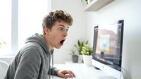 Young man with curly brown hair and light complexion, wearing gray hoodie, expressing shock while gaming on desktop computer in bright home office with plants.