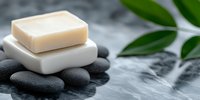 Natural soap bar on white ceramic holder with black spa stones and green leaves against gray marble background, wellness and body care concept.