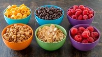 Colorful bowls filled with breakfast cereals, fresh raspberries, chocolate loops and cheese snacks arranged on dark wooden background, top view food photography.