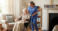 Professional caregiver assisting elderly woman in wheelchair at comfortable home living room. Healthcare support and senior care services concept.