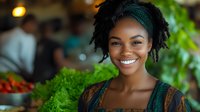 Young African American woman with natural hair and bright smile wearing green headband and ethnic pattern dress at local farmers market with fresh produce background.