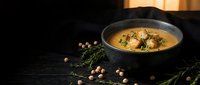 Creamy mushroom soup with herbs in rustic bowl on dark wooden table with scattered chickpeas and fresh rosemary sprigs for cozy dining atmosphere.