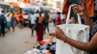Person of African descent holding white fabric bag at outdoor market with blurred crowd in background, suitable for retail, shopping, commerce, and lifestyle content.