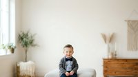 Young boy in formal attire sitting on bed in bright, minimalist bedroom with natural light, suitable for family, lifestyle, and children's content projects.