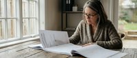 Middle-aged Caucasian woman with glasses reading documents at wooden table near window in bright home office space for business and professional work.