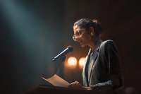 Professional woman in glasses speaking at podium with dramatic lighting, creating atmosphere of authority and importance during conference presentation.