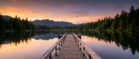Wooden dock extending into calm lake at sunrise with forested mountains and golden sky reflected in still water, suitable for travel and nature backgrounds.