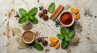 Aromatic spices and herbs arranged on textured stone surface including cinnamon sticks, star anise, turmeric, coriander seeds and fresh mint leaves for cooking.