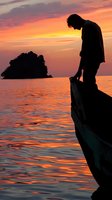 Man standing on wooden boat bow at sunset over calm ocean waters with distant island silhouette, golden hour seascape for travel and contemplation themes.