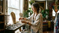 Female artisan with long curly hair arranging jewelry pieces at a wooden table in a bohemian studio filled with plants, textiles, and vintage decor. Handmade accessories workshop setting.