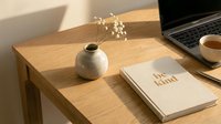 Cozy minimalist workspace with wooden desk, ceramic vase, dried flowers, be kind notebook, tea cup, and laptop in warm sunlight.