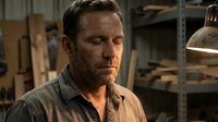 Mature Caucasian male craftsman with beard wearing work shirt in industrial workshop setting with tools and storage shelves in background.