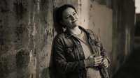 Young woman in denim jacket leaning against weathered concrete wall in urban setting with dramatic lighting and contemplative mood for lifestyle photography.