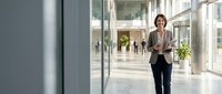 Professional middle-aged Caucasian businesswoman walking confidently through modern corporate office lobby with glass architecture and natural lighting.