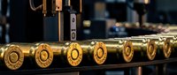 Brass ammunition cartridges lined up on industrial manufacturing conveyor belt in factory production line with machinery equipment in background.