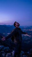 Young man with arms outstretched celebrating freedom on mountain peak at twilight with city lights below for adventure and success concepts.