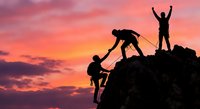 Silhouettes of three people climbing rocky mountain peak at sunset with dramatic pink and orange sky, teamwork and achievement concept for motivation.