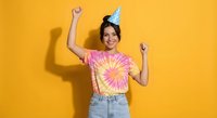 Young Hispanic woman celebrating birthday in colorful tie-dye shirt and party hat with raised fists showing excitement on bright yellow background.
