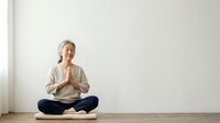 Mature woman with gray hair, Caucasian ethnicity, sitting in lotus position on wooden floor against white wall, hands in prayer pose at chest, wearing beige cardigan and jeans, practicing meditation or yoga mindfulness.
