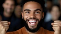 Young African American man celebrating success with raised fists and bright smile in crowded environment. Expresses joy, achievement and positive energy.