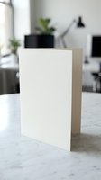 Blank white greeting card standing on marble surface in modern office interior, mockup template for custom design and branding.