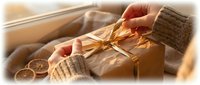 Hands wrapping kraft paper gift box with golden ribbon near dried citrus slices and twine for holiday present packaging and seasonal celebrations.