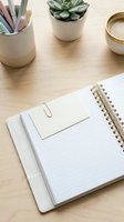 Open spiral notebook with gold paper clip and blank card on wooden desk, succulent plant and pastel pens in background, flat lay.