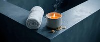 Rolled white towel and lit candle with scattered stones on geometric concrete surface for spa and wellness design projects.
