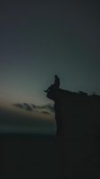 Silhouette of person sitting alone on cliff edge against moody dark sky at dusk, contemplative solitary figure overlooking vast landscape in atmospheric evening light.
