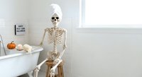 Skeleton sitting on wooden stool in modern bathroom with pumpkins and candles for Halloween decoration and spooky autumn interior design concept.