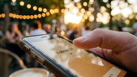 Hand holding smartphone with Follow Us message on screen at outdoor cafe with bokeh lights at sunset.