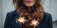 Young woman in dark winter coat with fur collar holding glowing sparklers creating warm festive atmosphere for holiday celebrations and winter fashion.