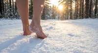 Bare feet walking through fresh snow in winter forest with golden sunlight filtering through trees creating magical cold therapy wellness moment.