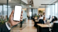 Hand holding smartphone with blank screen in modern office environment with colleagues working at desks in background. For app mockups and business presentations.