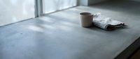 Minimalist concrete countertop with ceramic mug and folded towel in modern kitchen interior with natural window light creating soft shadows.