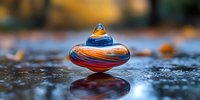 Colorful glass spinning top with swirling orange and blue patterns reflects on wet surface against blurred autumn background, creating artistic macro composition.