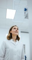 Young woman with shoulder-length brown hair, Caucasian ethnicity, wearing white lab coat, looking upward at digital interface display in modern laboratory or medical facility setting.